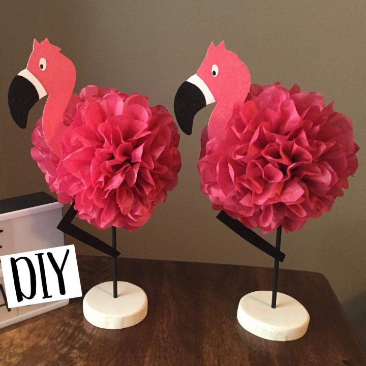 Two handmade flamingo centerpieces with hot pink tissue paper pom poms stand next to a sign that says, "DIY".