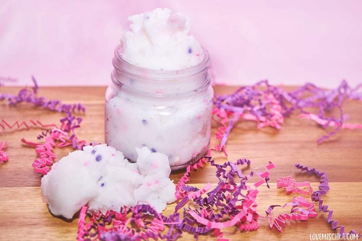 Fluffy white sugar scrub with pink and white sprinkles in a mason jar on a piece of wood with pink and purple confetti.