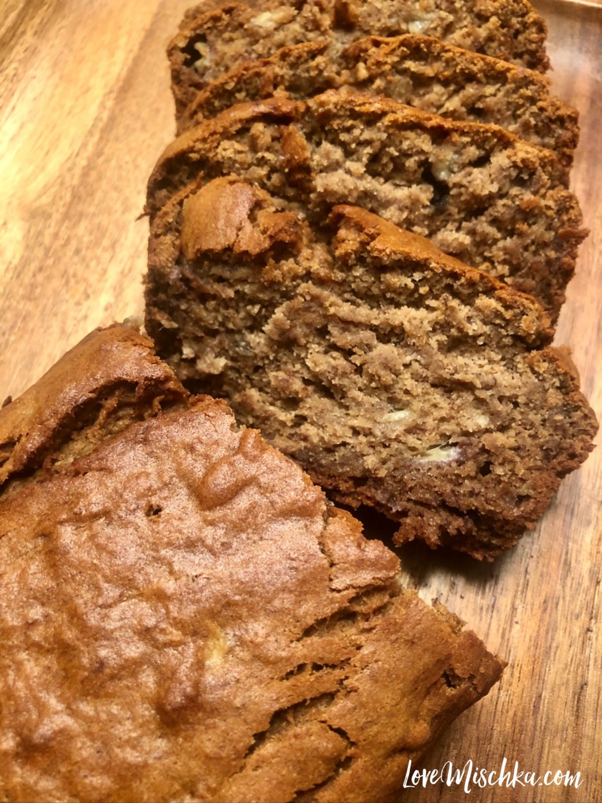 A brown loaf of moist banana bread with a few pieces sliced and laid out on wood.