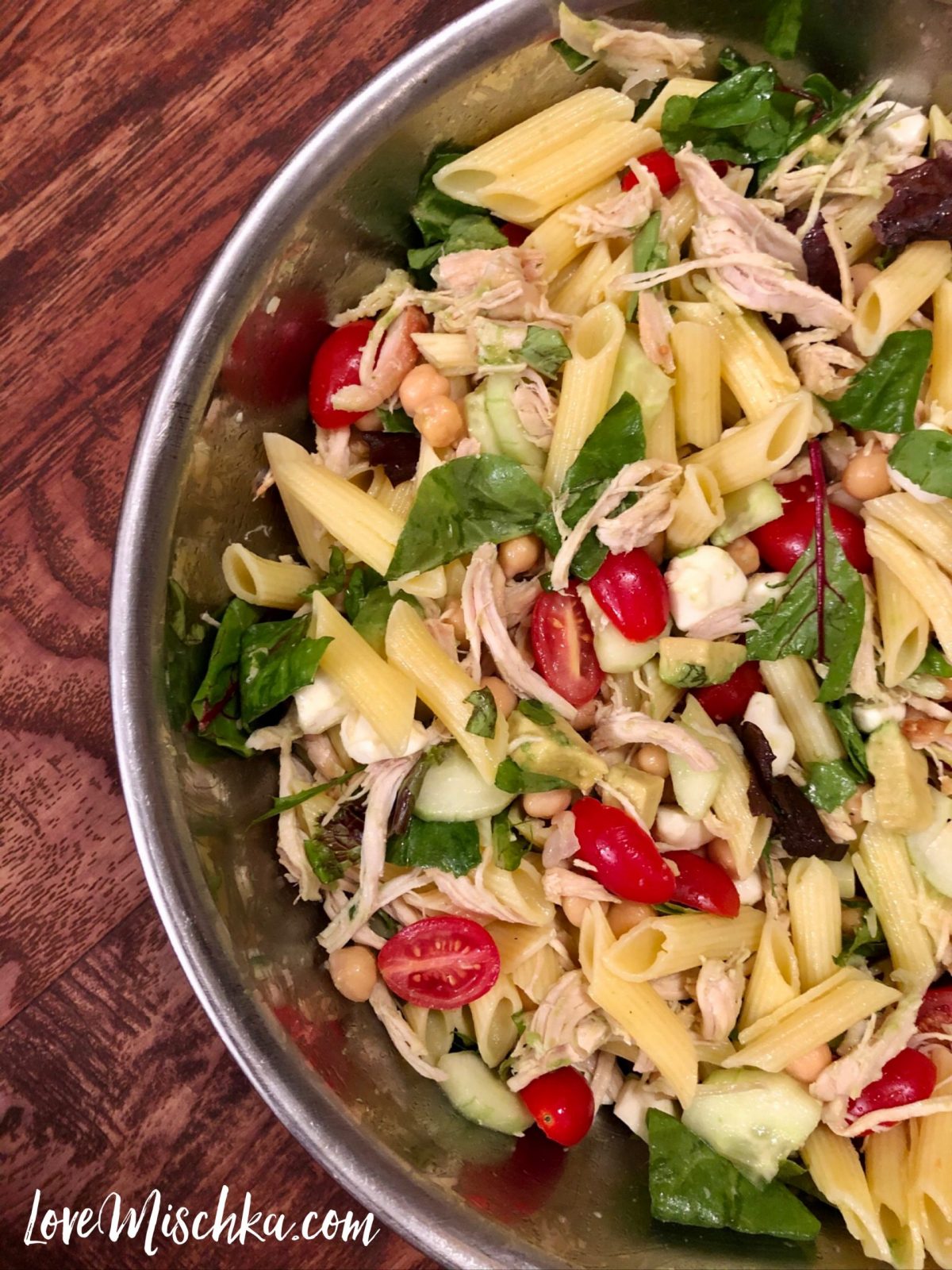 A bowl of pasta salad - pasta, chicken, red tomatoes, spring mix lettuce, green basil, tan chickpeas, and more.