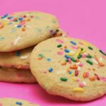 A stack of tan, round cookies with bright rainbow sprinkles on a neon pink table.