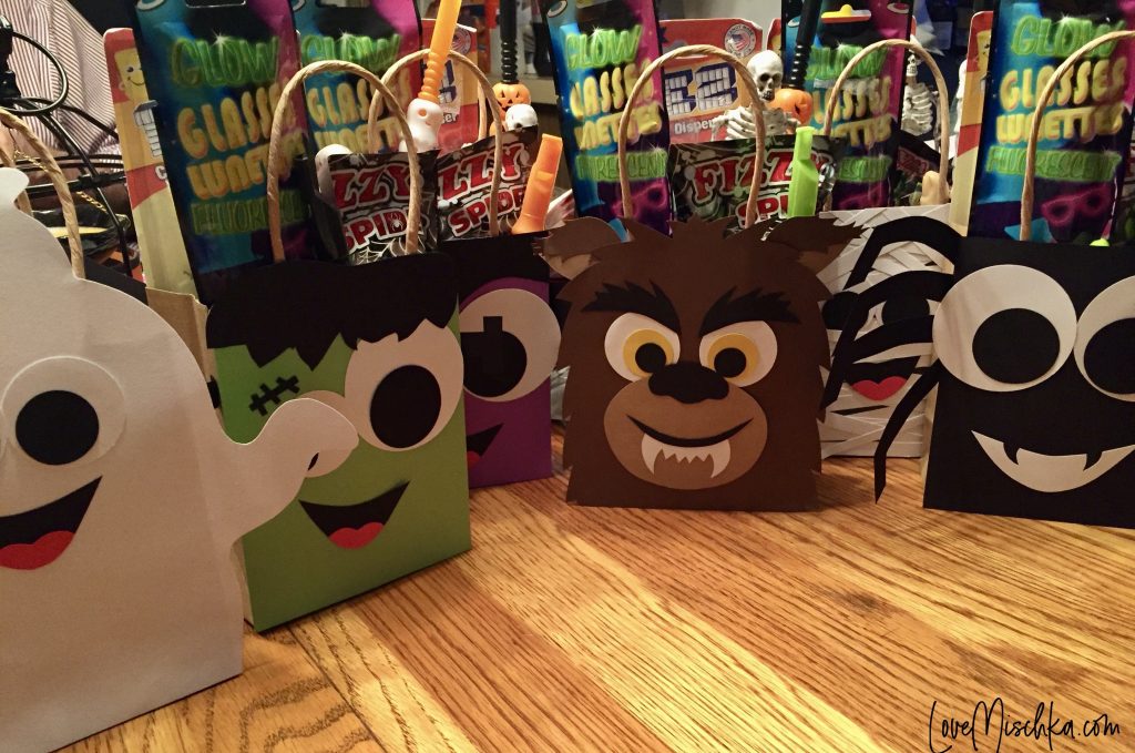 A white ghost goodie bag, a green and black Frankenstein bag, a purple and black bag, a brown werewolf bag with yellow eyes and sharp teeth, and a black spider bag. Each goodie bag is filled with candy and Halloween toys.