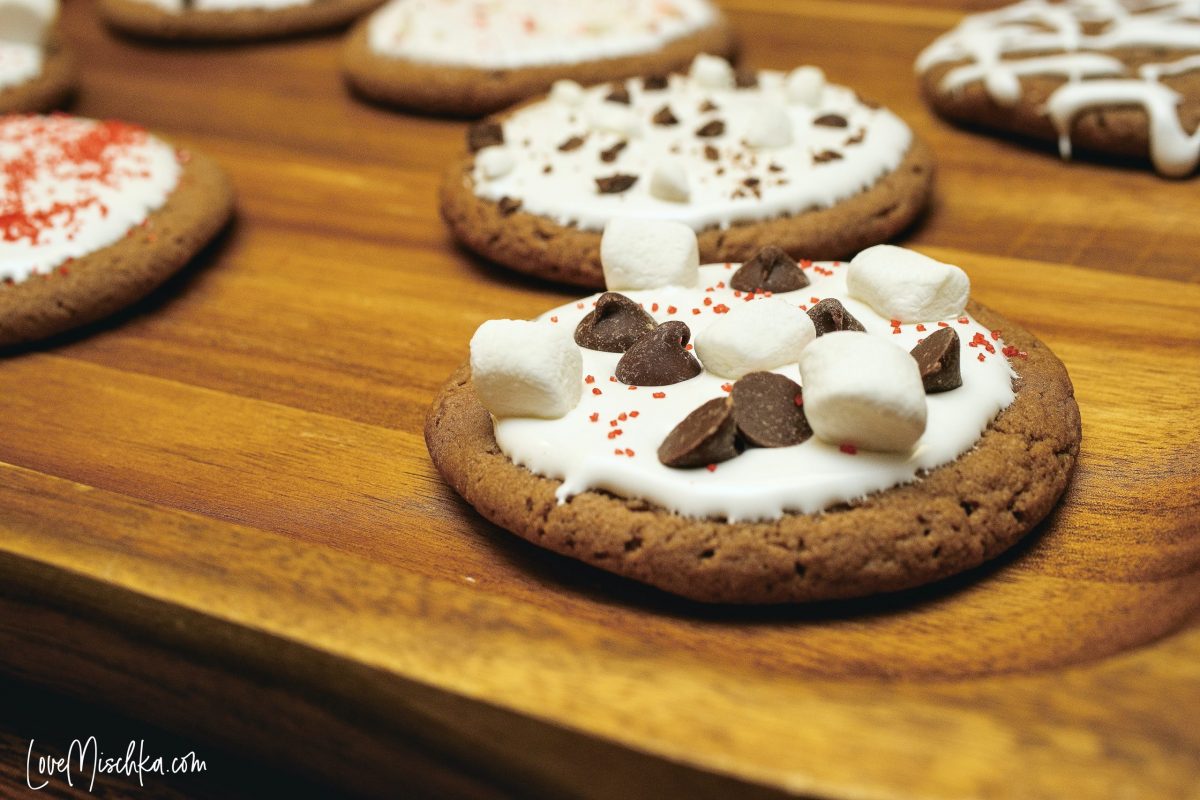 Round, dark brown hot chocolate cookies topped with marshmallow creme, marshmallows, chocolate, and red sprinkles on a wooden serving tray.