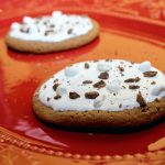 Two dark brown hot chocolate cookies topped with white icing, chopped chocolate, and chopped marshmallows.