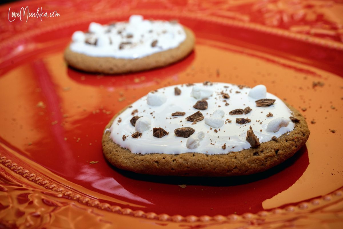 Two dark brown hot chocolate cookies topped with white icing, chopped chocolate, and chopped marshmallows.