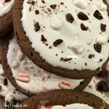 Close Up of a stack of dark brown hot chocolate cookies with marshmallow creme icing and various toppings - marshmallows, candy canes, chopped chocolate.