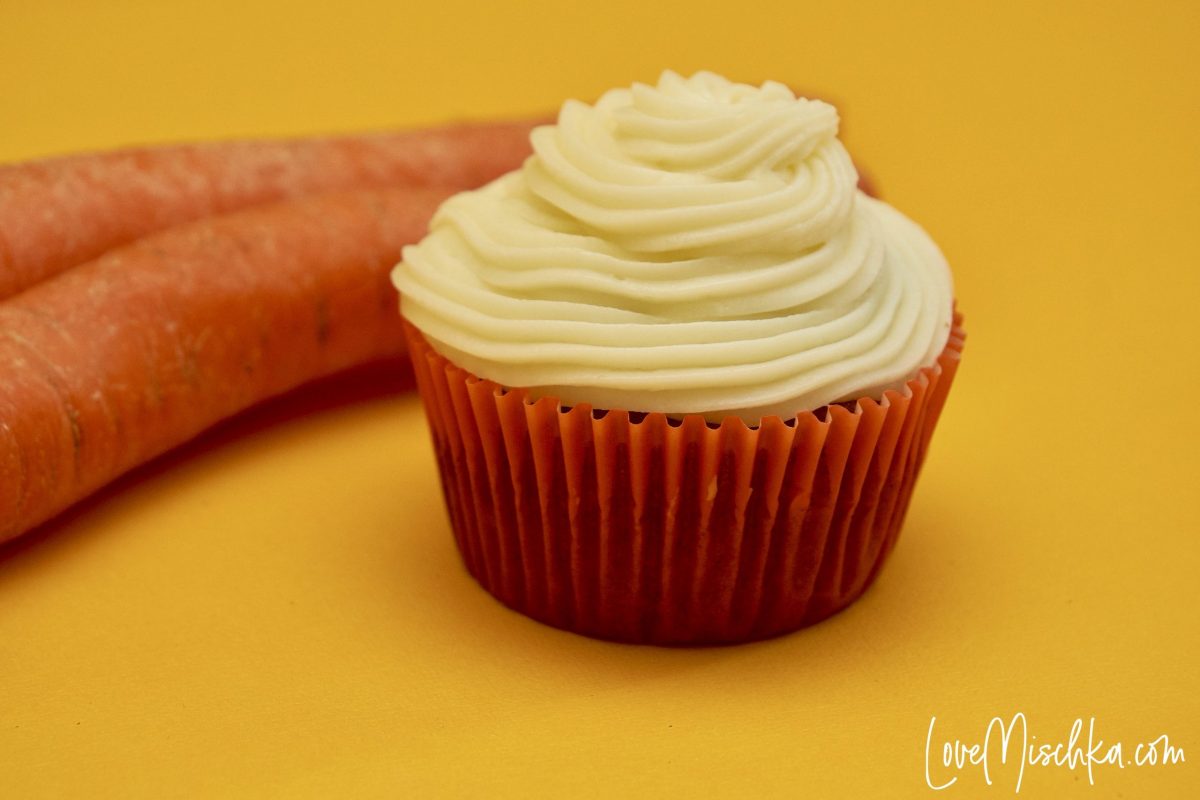One Carrot Cake Cupcake with Cream Cheese Frosting with an orange wrapper and two carrots next to it.