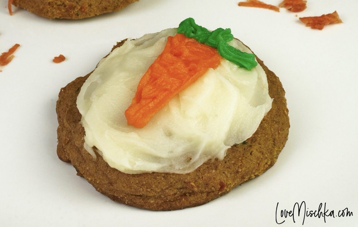 A round carrot cookie with white frosting and an adorable carrot design on a white plate with carrot shavings.