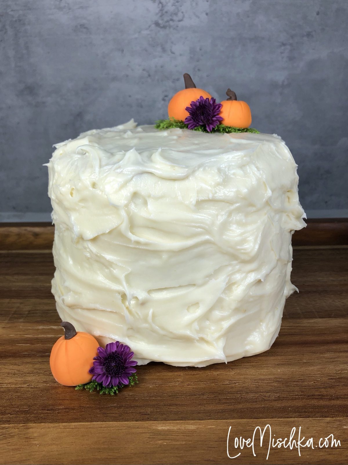 A tall Cake with fluffy frosting with three small pumpkins and two purple flowers.