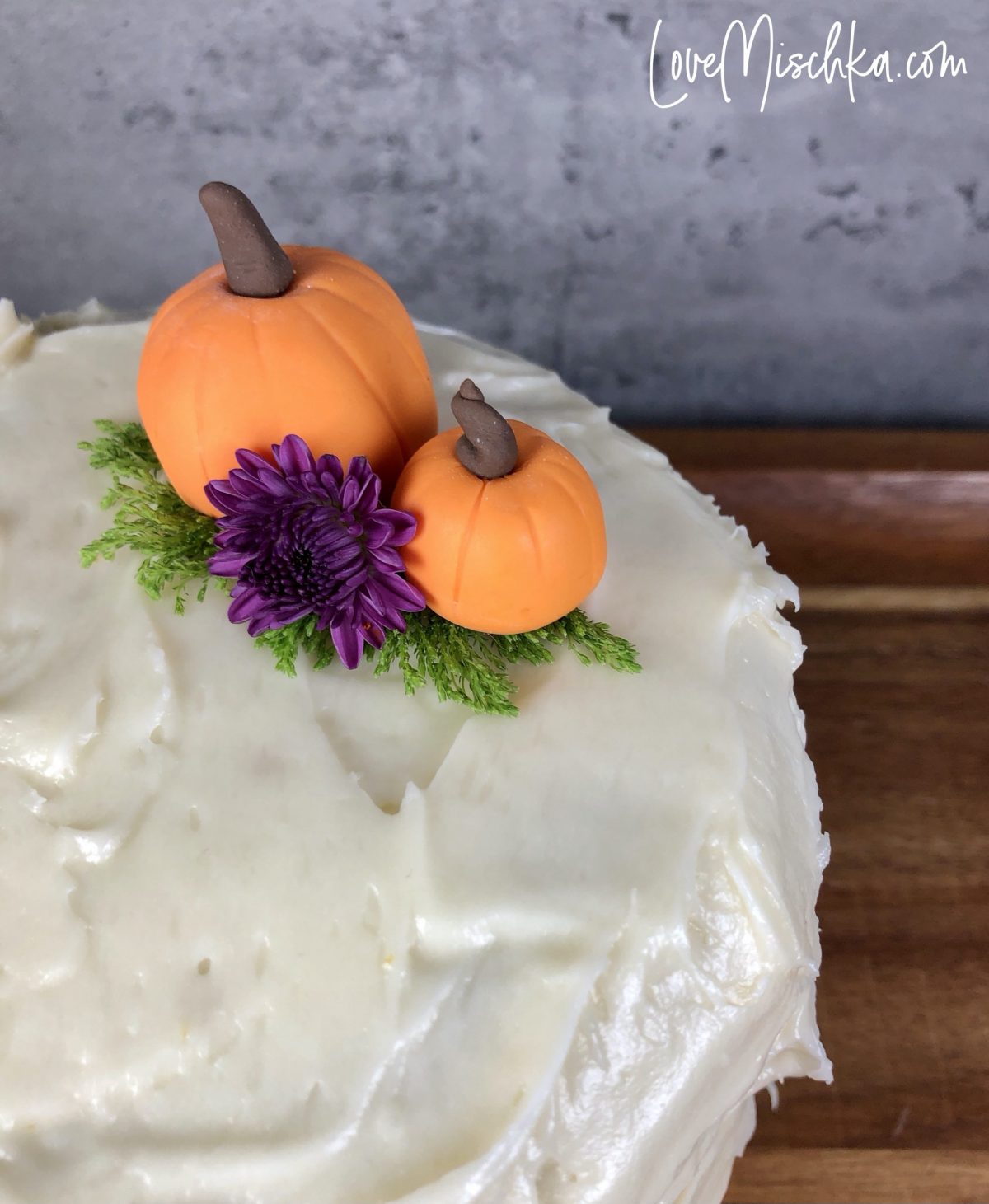 Round cake with fluffy cream cheese frosting, two cute pumpkins, and a purple flower on a wooden platter.