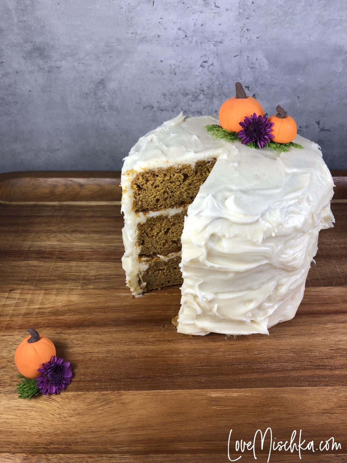 Tall Pumpkin Cake with a Slice Missing. Two Pumpkins on the cake and one on the wooden serving tray.