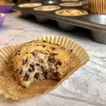 A banana chocolate chip muffins sits on a table in front of a silver baking tin of other banana muffins. A big bite is taken out so we can see all the chocolate inside the moist muffin.
