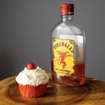 A cupcake with fluffy white frosting and a fireball on top sits next to a half full bottle of Fireball Whisky.