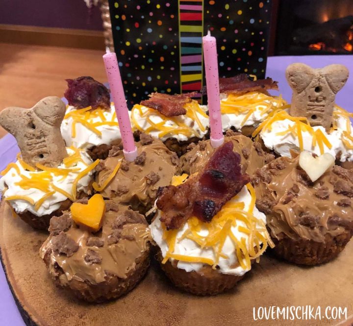 Small pupcakes covered in peanut butter, cheese, bacon, and bones sit on a wooden board with two pink candles.