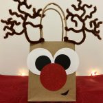 A brown kraft bag with big cartoon eyes, a red glitter circular nose, and a crooked smile. The Reindeer also has complex antlers made out of brown pipe cleaner.