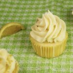 A pale yellow lemon cupcake with beautiful swirled vanilla buttercream sits on a neon green and white plaid cloth next to a slice of lemon.