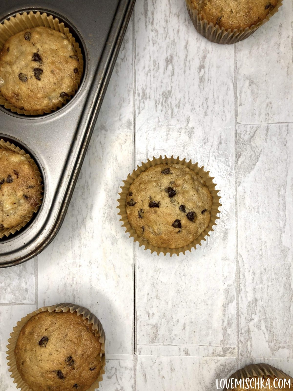 Four Golden Banana Bread Chocolate Chip Muffins are on a white, marble table with a stainless steel baking pan full of muffins next to them.