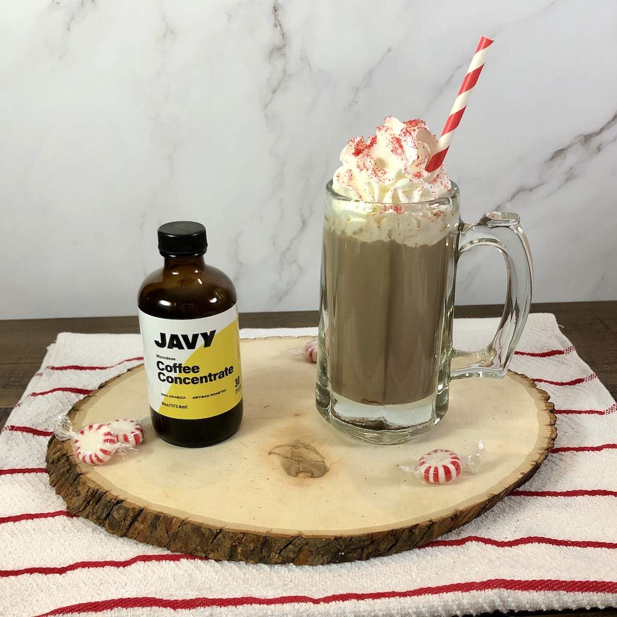 A bottle of Javy Coffee Concentrate sits next to a mug of homemade Peppermint Mocha topped with whipped cream, red sanding sugar, and a red and white striped straw.