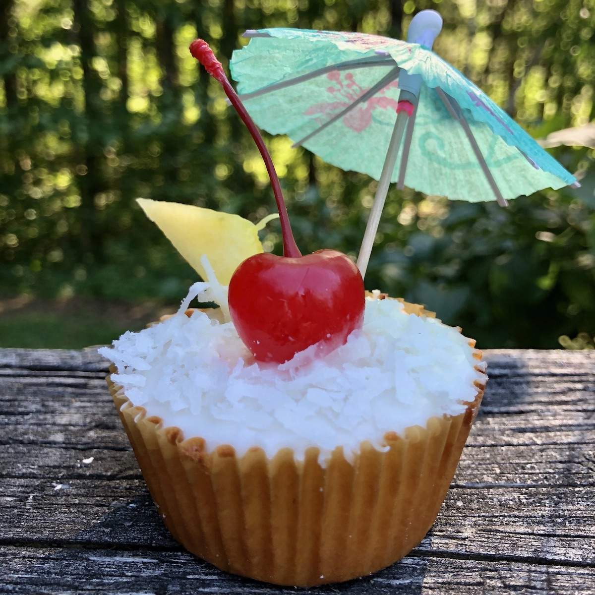 A golden cupcake topped with white frosting, coconut flakes, a bright red cherry, a fresh piece of pineapple, and a mint green cocktail umbrella.