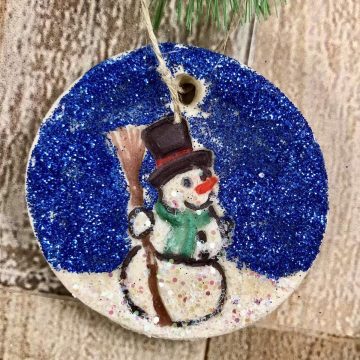 A homemade salt dough ornament in the shape of a circle. A snowman is imprinted in the middle and colored with marker and glitter.
