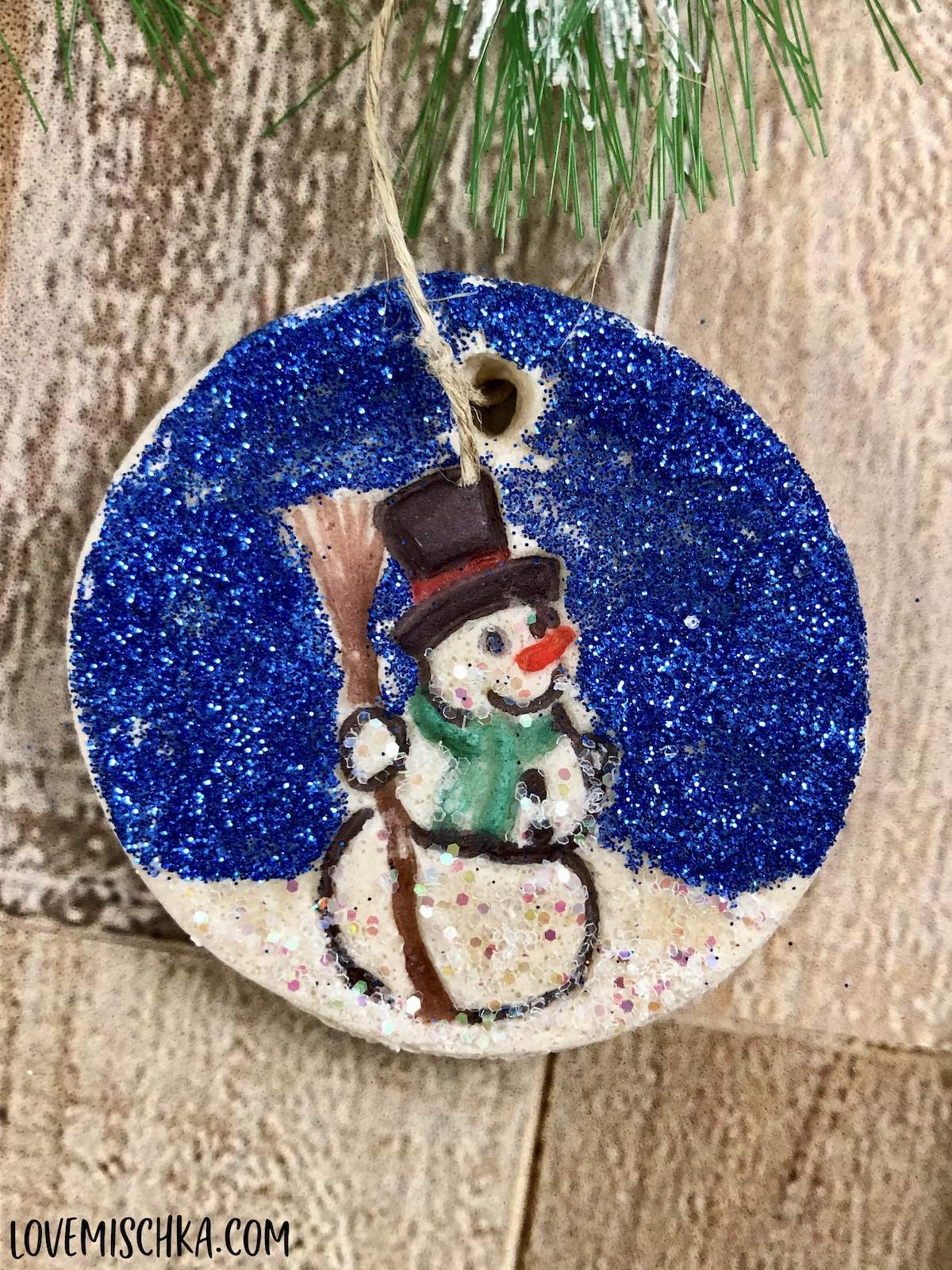 A homemade salt dough ornament in the shape of a circle. A snowman is imprinted in the middle and colored with marker and glitter.