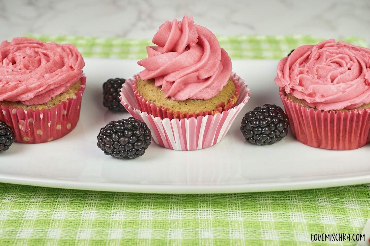 Three Earl Grey Cupcakes topped with hot pink, blackberry buttercream frosting sit on a plate with fresh blackberries.