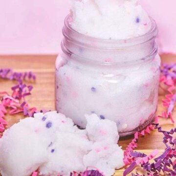 A mason jar full of white, fluffy homemade body scrub with pink and purple confetti in and surrounding it.