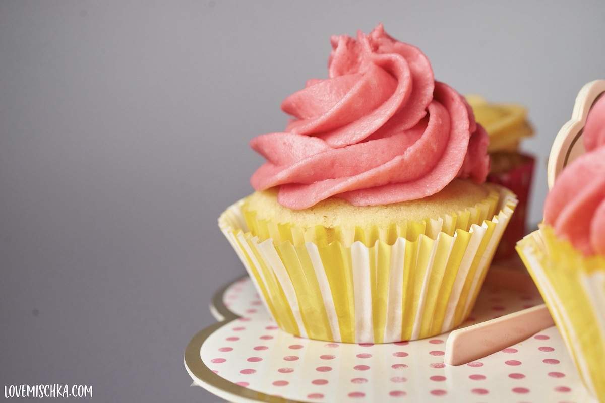 A yellow lemon cupcake in a yellow and white striped cupcake liner topped with pink blackberry buttercream frosting in a swirl.