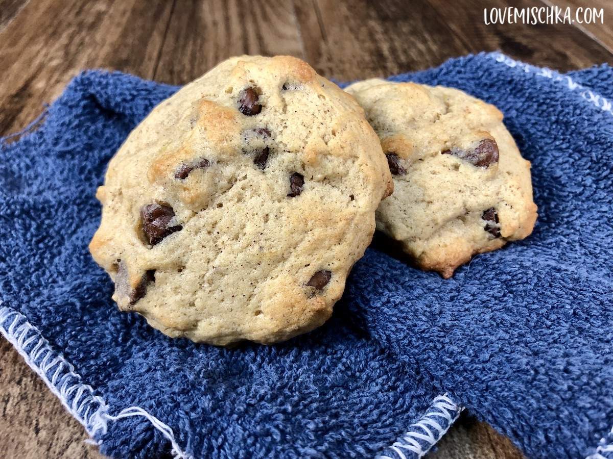 Two tan banana bread cookies with brown chocolate chips on a dark blue cloth.