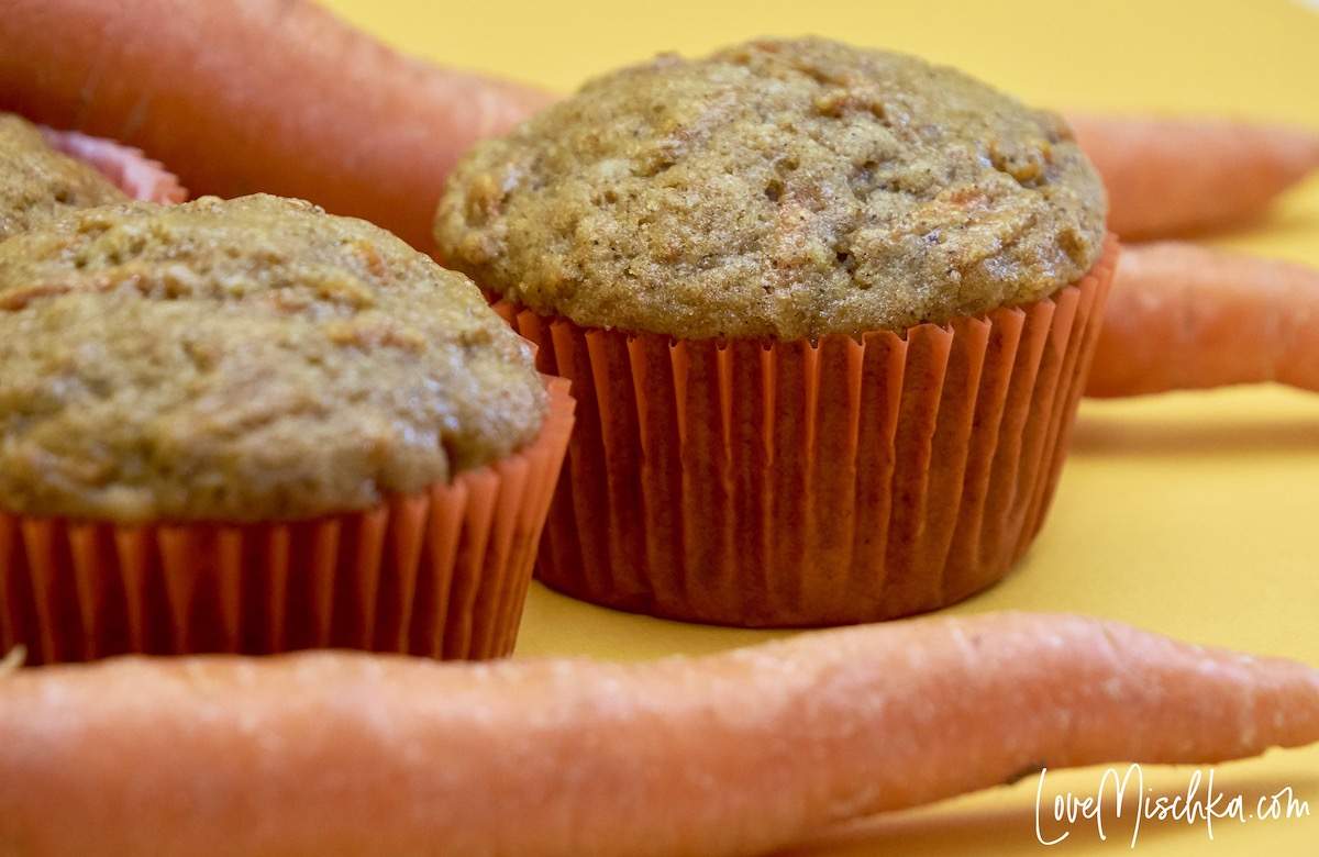 Brown Carrot Cake Muffins in orange cupcake liners surrounded by fresh, whole carrots.