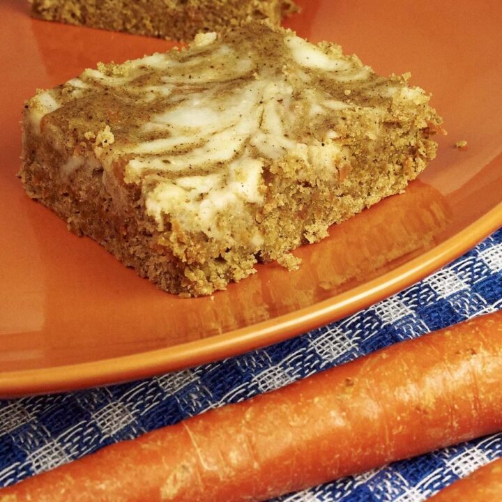 A tan carrot cake bar swirled with white cream cheese sits on an orange plate next to orange, whole carrots on a blue and white plaid cloth.