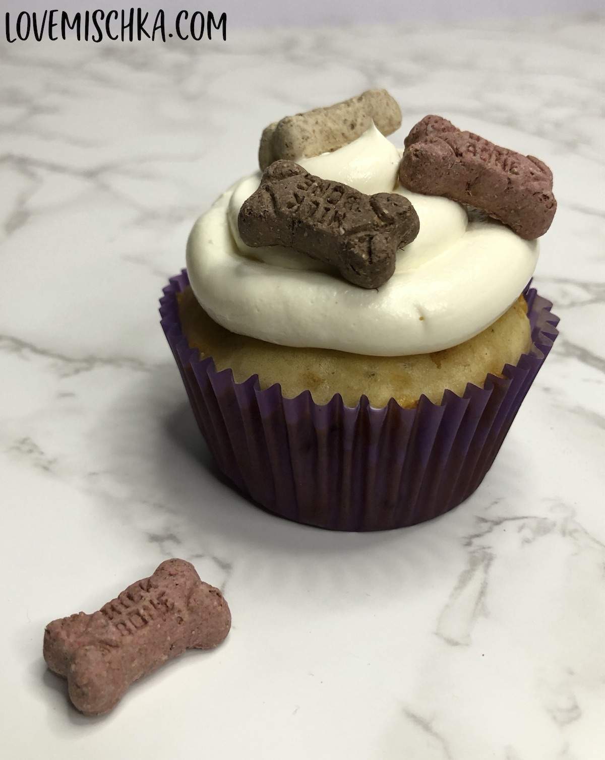 A dog cupcake in a dark purple cupcake wrapper, topped with white cream cheese and three mini milkbones - red, dark brown, and tan. 