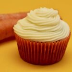 One Carrot Cake Cupcake in a dark orange wrapper, topped with silk cream cheese frosting next to two whole carrots.