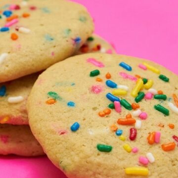 A stack of round, golden funfetti cookies with bright rainbow sprinkles with a hot pink background.