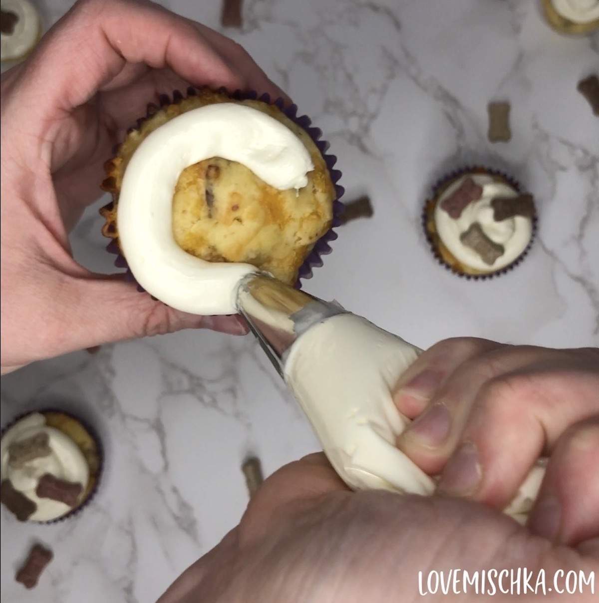 A hand pipes white cream cheese frosting on a golden bacon cheddar cupcake with a piping bag and silver piping tip.