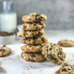 A stack of golden brown Almond Joy Cookies next to coconut chocolate chip cookies, a white bowl of milk chocolate chips, and a glass of white milk.