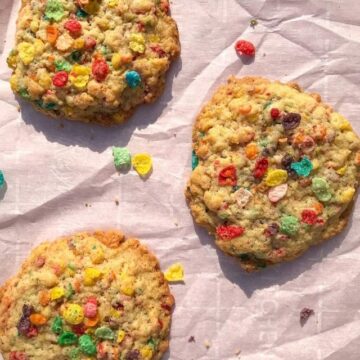 Three bright Fruity Pebbles Cookies with a rainbow of colors sit on a parchment-lined, pink baking sheet.