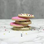 Round Sugar Cookies topped with pink and white buttercream frosting for cookies are stacked on top of one another and surrounded by rainbow sprinkles.