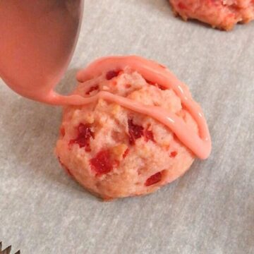 Light pink cherry glaze is drizzled on a pink maraschino cherry cookie that has bright red pieces of maraschino cherries in it.