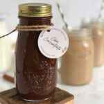 A glass bottle full of reddish brown chai concentrate made from tea bags with two iced chai lattes in the background.