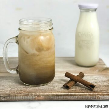 A brown, tan, and white swirl iced chai tea latte sits on a light brown wooden plank next to two sticks of dark brown cinnamon and a milk bottle of white milk.