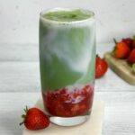 A tall glass with a swirl of white milk and green matcha green tea on top of red strawberry puree on a white marble coaster with a fresh strawberry next to it.