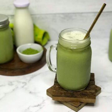 A vibrant green iced matcha latte with a gold straw sits next to a milk bottle of white milk, a small white bowl or bright green matcha powder, and another vibrant green tea latte in a mason jar.