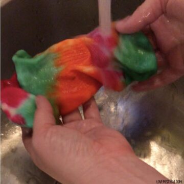 A newly tie-dyed sock that is neon green, magenta, and orange is held under a stream of water.