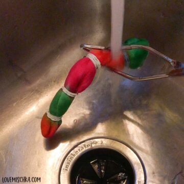 A magenta, green, and orange sock that is wrapped with rubber bands is held under a faucet while water runs through the sock and down the stainless steel drain.