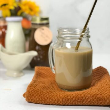 A mason jar glass full of tan pumpkin spice chai latte on an orange dish towel with a gold straw. The three ingredients - milk, pumpkin sauce, and chai concentrate - sit in the back in bottles.