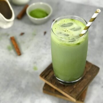 A bright green matcha chai latte in a glass on top of dark brown wooden coasters next to the ingredients of this matcha chai latte recipe - chai concentrate, green tea powder, and milk.