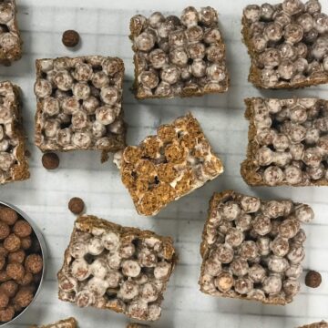 Several square-shaped, brown cocoa puff treats lay on a white sheet of parchment paper. One Cocoa Puff Bar is on it's side showing it's thickness and the crispy inside of the favorite cereal.