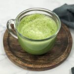 A glass mug of a bright green, hot matcha latte with a frothed layer on top. The mug sits on a circular, dark brown, wooden serving tray.