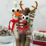 A brown, milk chocolate Reindeer Pretzel Rod with golden, pretzel antlers and a bright red licorice bow and a white chocolate dipped Snowman Pretzel rod with a bright orange mike and ike candy nose and a bright red licorice scarf stand in a decorative glass vase with other Christmas Pretzel Rods.
